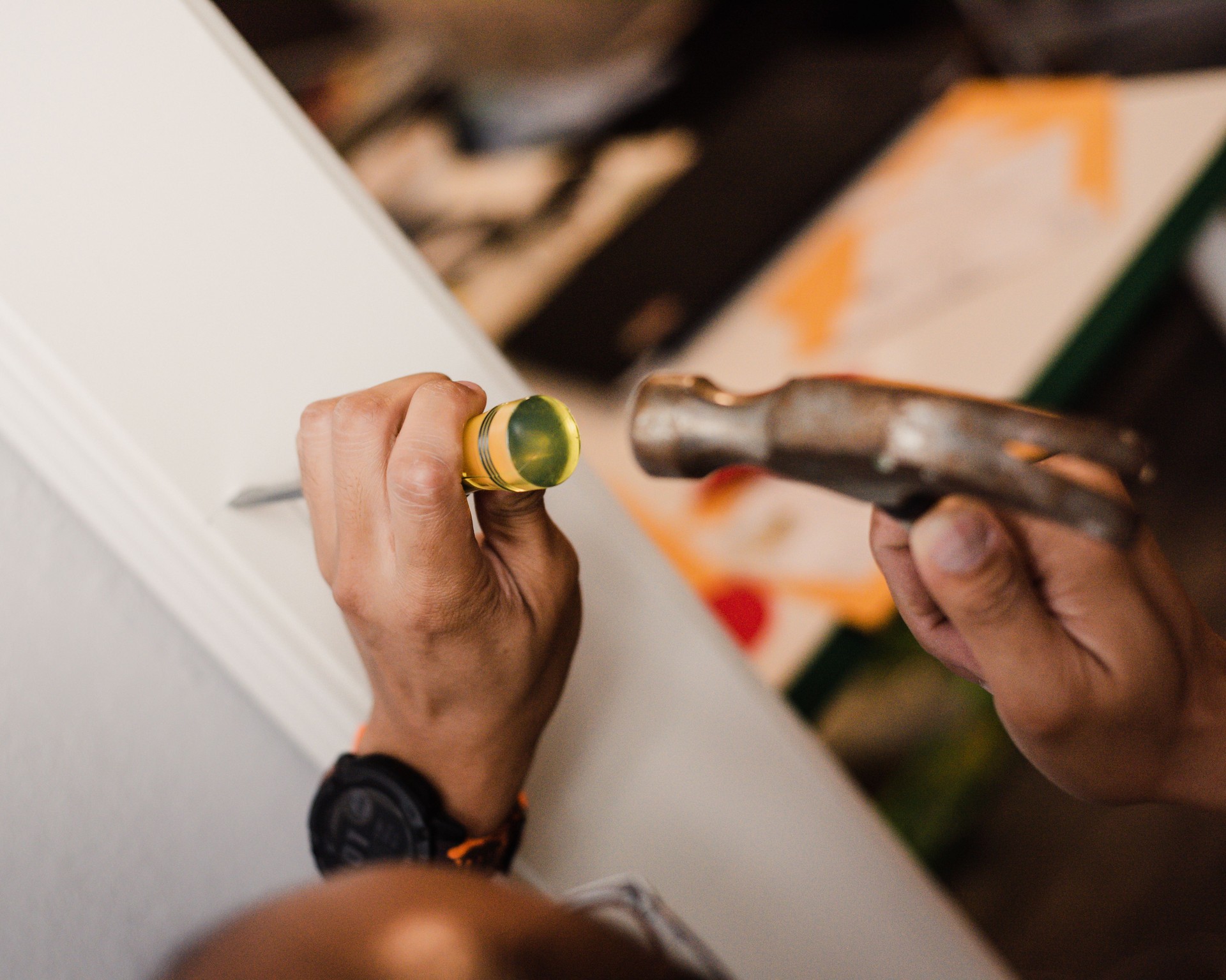 Panorama view chisels hinge recess into white door frame with hammer by Asian handyman. Digital smartwatch visible. Background includes blurred table with yellow object. DIY task shows precision